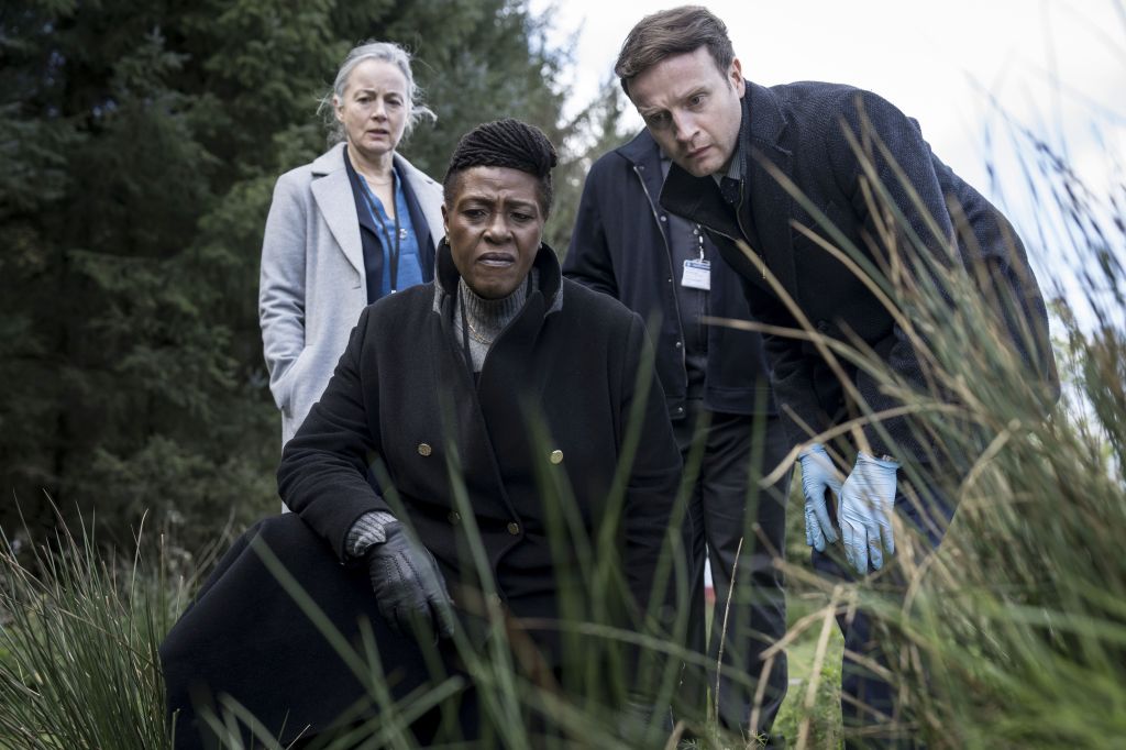 Ellis und Harper stehen im hohen Gras und schauen auf einen Tatort, der außerhalb des Fotos ist.